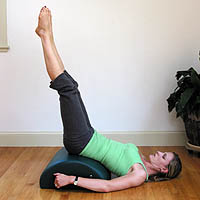 A woman lying on her back with a mini barrell beneath her lower back, raising her legs straight up